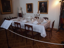 Ruokapöytä, jolla on aatelisvaakunoilla koristettu astiasto, lasisto ja ruokailuvälineet.